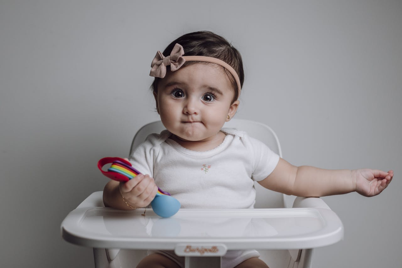 Cute baby girl sitting in a high chair holding a colorful toy, looking curious and innocent.