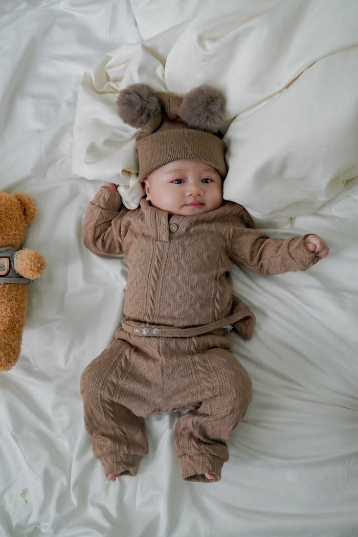 A cute baby lying on a white bed wearing a brown outfit and hat with pom-poms.